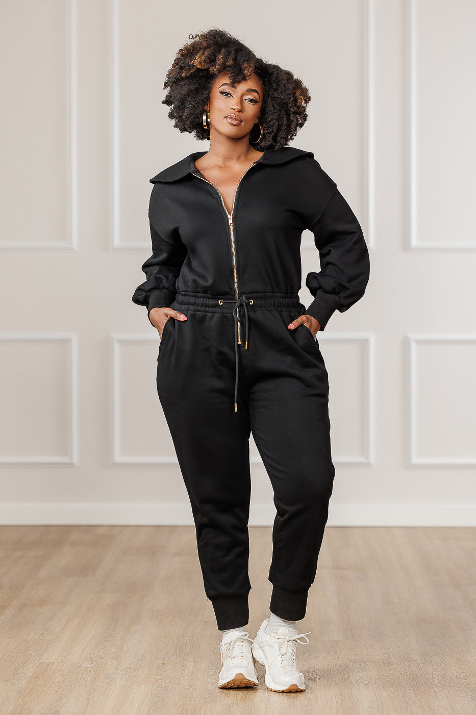 A woman standing in a room with light-colored walls wearing a black Sutton Puffed Sleeve Jumpsuit with an open zipper front and collar neckline, paired with white sneakers.