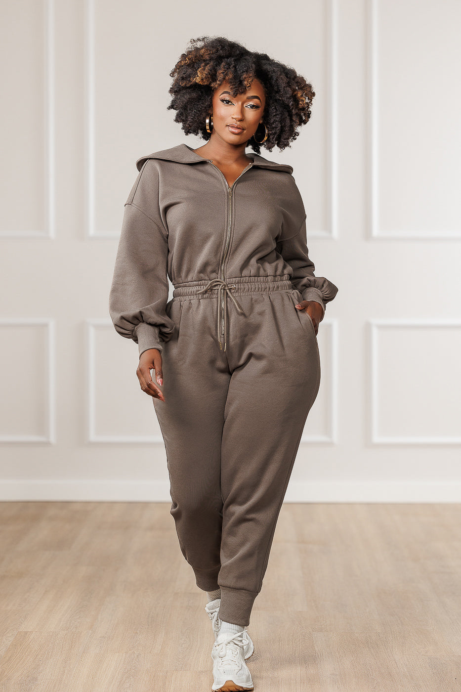 A model wearing a brown Sutton Puffed Sleeve Jumpsuit with a collar neckline, open zipper front, and elasticized waistband.